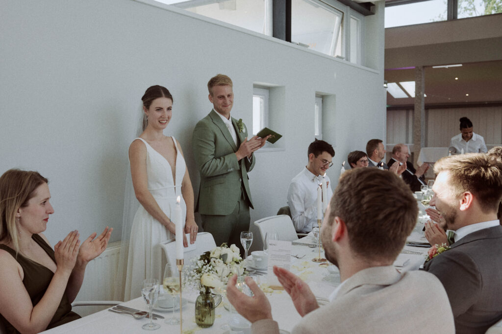 Braut und Bräutigam stehen während einer Hochzeitsfeier in Bielefeld an einem Tisch. Die Braut trägt ein weißes Kleid und der Bräutigam einen hellgrünen Anzug. Beide lächeln. Die Gäste sitzen um den Tisch herum und klatschen. Der Raum hat weiße Wände und große Fenster.