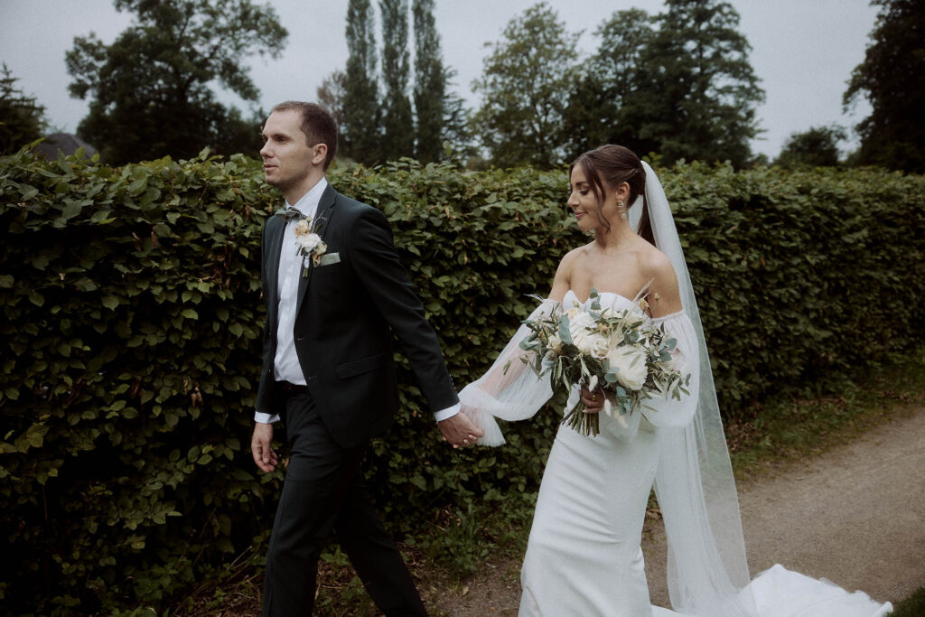 Braut und Bräutigam gehen Hand in Hand einen Feldweg in Bielefeld entlang. Die Braut trägt ein weißes Hochzeitskleid und einen Schleier und hält einen Brautstrauß mit weißen Blumen in der Hand. Der Bräutigam trägt einen dunklen Anzug und eine Ansteckblume. Hinter ihnen ist eine Hecke mit hohen Bäumen im Hintergrund.