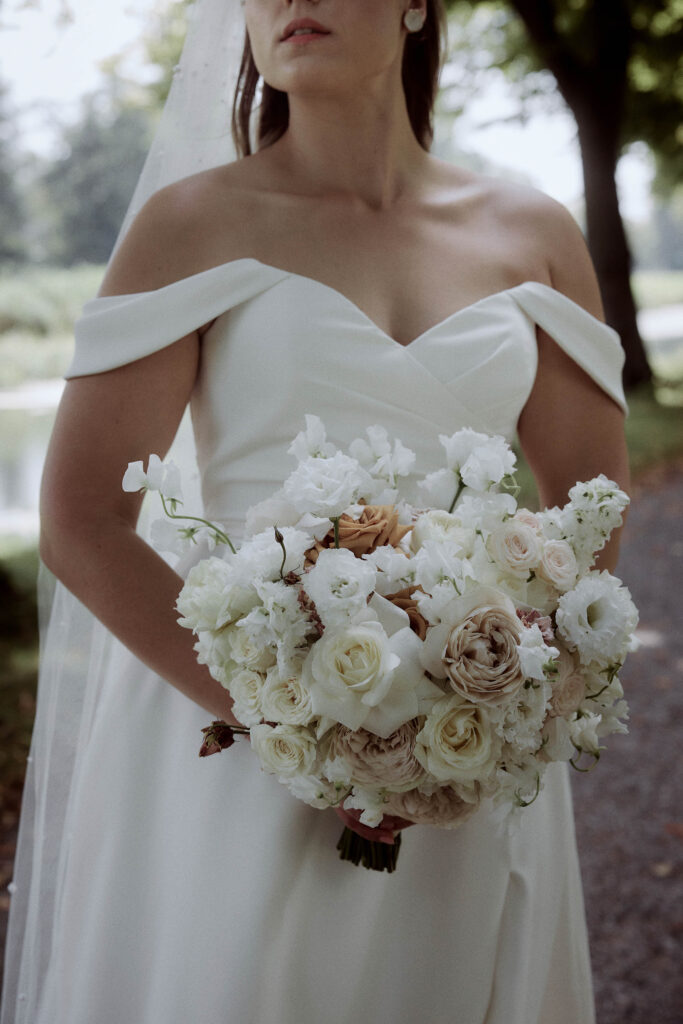 Braut in einem schulterfreien weißen Kleid mit einem Strauß weißer und rötlicher Blumen. Sie steht im Freien mit Bäumen und einem Gehweg im Hintergrund.