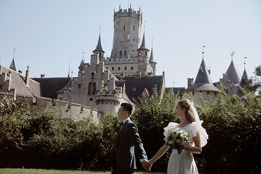 Ein Paar in formeller Kleidung hält sich im Freien an den Händen, im Hintergrund das historische Schloss Marienburg. Die Türme und der zentrale Turm des Schlosses erheben sich majestätisch, während die Braut einen Blumenstrauß in den Händen hält, perfekt für ein Hochzeitsshooting. Die Umgebung ist hell und sonnig und verleiht diesem malerischen Moment Charme.