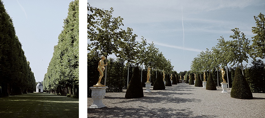 Das Bild zeigt die Herrenhäuser Gärten in Hannover, einen formellen Garten mit zwei Abschnitten. Links führt ein von hohen, gestutzten Bäumen gesäumter Grasweg zu einem kleinen Gebäude. Rechts ist ein Kiesweg zu sehen, der von Formschnittbäumen und goldenen Statuen auf Sockeln unter einem klaren Himmel gesäumt ist – ideal für eine standesamtliche Hochzeit.