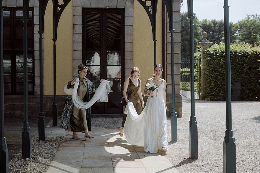 Eine Braut in einem weißen Kleid hält einen Blumenstrauß und geht unter einer dekorativen Struktur in den Herrenhäuser Gärten Hannover hindurch. Zwei Frauen begleiten sie, eine richtet die Schleppe der Braut. Die Kulisse umfasst einen gewölbten Eingang und üppiges Grün, perfekt für eine emotionale Trauung.