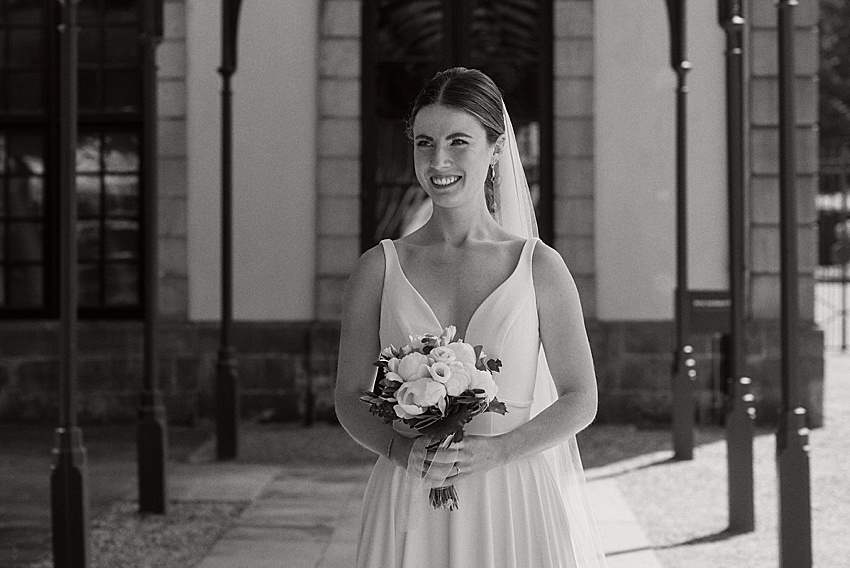 Eine Braut in einem ärmellosen weißen Brautkleid steht draußen, lächelt und hält einen Blumenstrauß bei ihrer standesamtlichen Hochzeit. Sie befindet sich unter einer Reihe von Bögen in den wunderschönen Herrenhäuser Gärten in Hannover, mit einem Gebäude im schwarz-weißen Hintergrund.