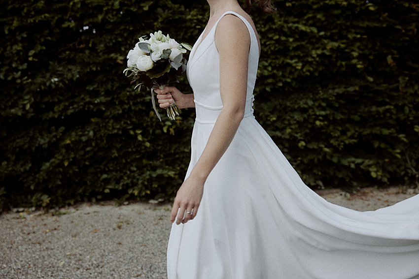 In einem fließenden weißen ärmellosen Kleid geht eine Brautanmutig den Kiesweg entlang und hält einen Strauß weißer Blumen und Grünpflanzen in der Hand. Das üppige Laub der Herrenhäuser Gärten in Hannover bietet eine bezaubernde Kulisse für diese standesamtliche Hochzeit.