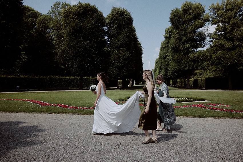 Bei einer standesamtlichen Hochzeit in Hannover geht eine Braut in einem weißen Kleid einen Kiesweg in den Herrenhäuser Gärten entlang. Zwei Frauen folgen ihr und tragen ihre Schleppe, umgeben von gepflegten Rasenflächen, leuchtend roten Blumen und hohen Bäumen unter dem wolkigen Himmel.