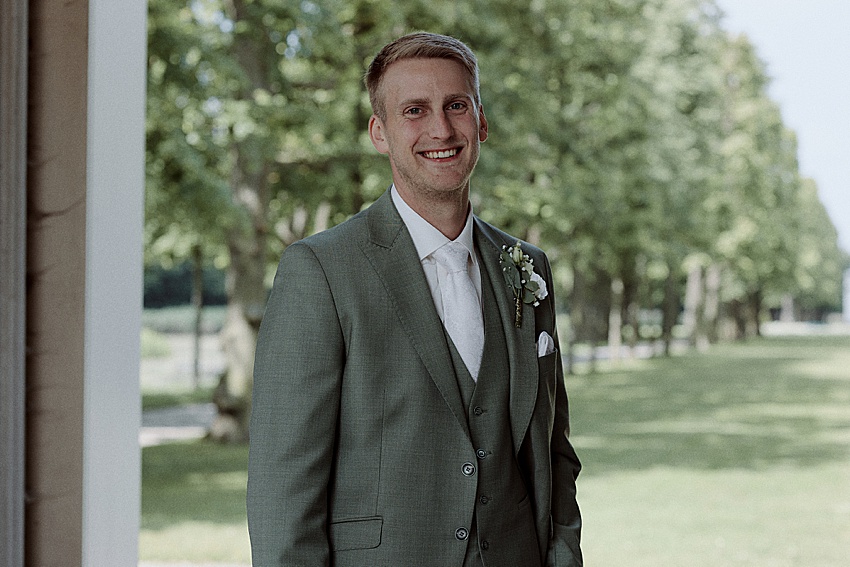 Ein Mann in einem grünen Anzug mit weißem Hemd und Krawatte steht vor dem Veranstaltungsort der Standesamtlichen Hochzeit und lächelt, während er die Ansteckblume an seinem Revers zurechtrückt. Bäume und Gras der Herrenhäuser Gärten Hannover sind unter einem klaren Himmel zu sehen und verleihen diesem Moment Charme.