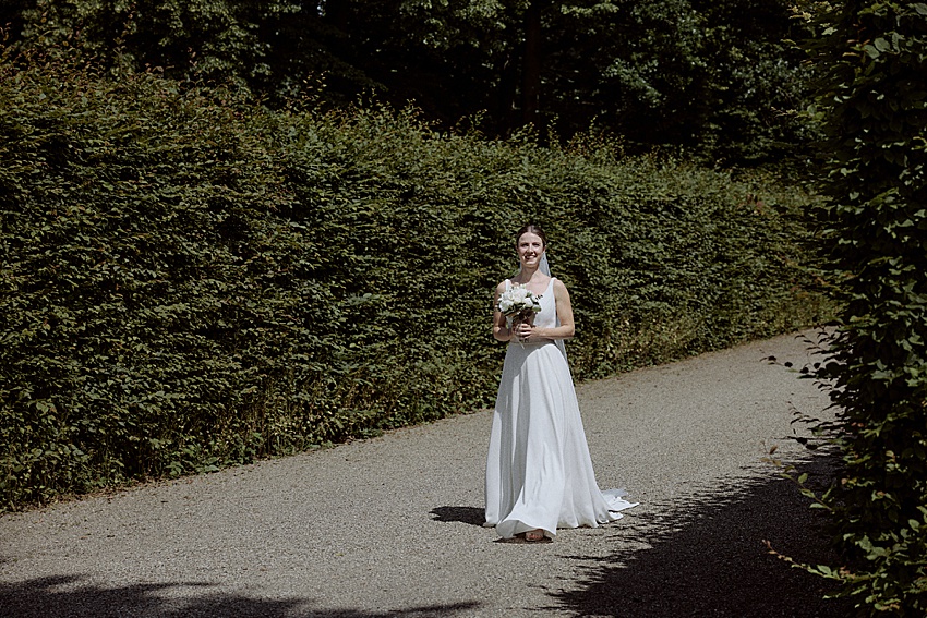 Eine Braut in einem weißen Kleid steht auf einem Kiesweg und hält einen Blumenstrauß in der Hand. Sie ist umgeben von hohen grünen Hecken unter dem teilweise bewölkten Himmel in den Herrenhäuser Gärten Hannover.