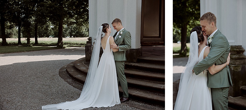 Braut und Bräutigam stehen dicht beieinander auf Steinstufen neben einem Gebäude und halten einen zärtlichen Moment während ihres First Looks an ihrer Hochzeit fest. Das weiße Kleid der Braut mit dem langen Schleier bildet einen schönen Kontrast zum grünen Anzug des Bräutigams, während sie sich vor der üppigen Kulisse der Herrenhäuser Gärten in Hannover umarmen.