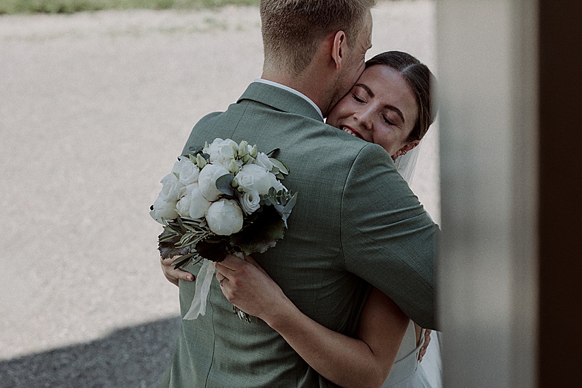 Ein Brautpaar umarmt sich herzlich vor der Standesamtlichen Hochzeit. Die Frau lächelt und hält einen Strauß weißer Blumen in der Hand. Der Mann trägt einen grünen Anzug. Im Hintergrund sind Kies und Grün zu sehen, die an die bezaubernden Herrenhäuser Gärten in Hannover erinnern.