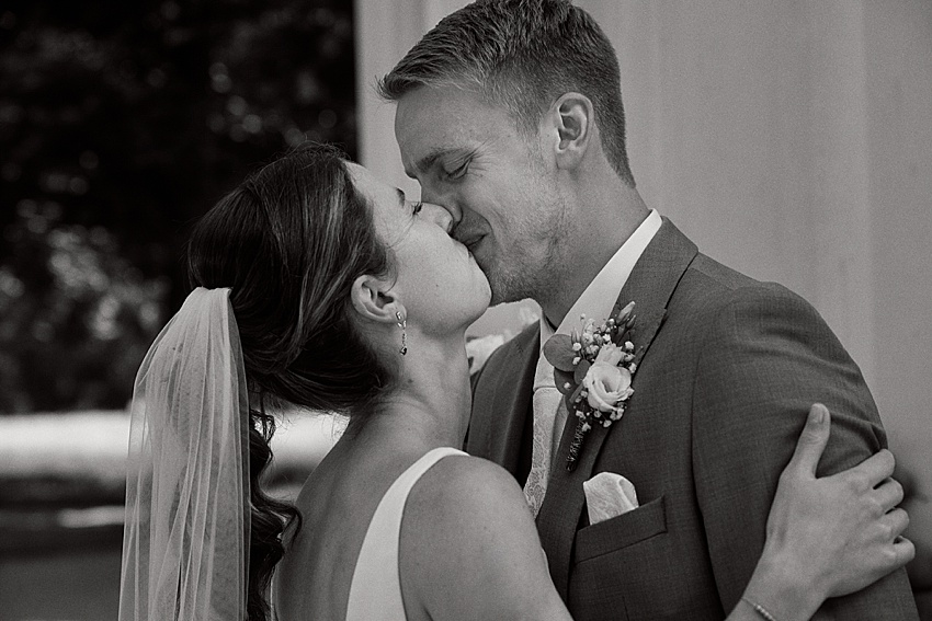 Auf einem zeitlosen Schwarzweißfoto küssen sich Braut und Bräutigam während ihres First Looks vor ihrer standesamtlichen Hochzeit zärtlich. Die anmutig verschleierte Braut umarmt ihren Bräutigam im Anzug vor der sanft verschwommenen Kulisse des Charmes Hannovers. Beide haben einen entspannten Gesichtsausdruck und fangen einen intimen Moment ein.