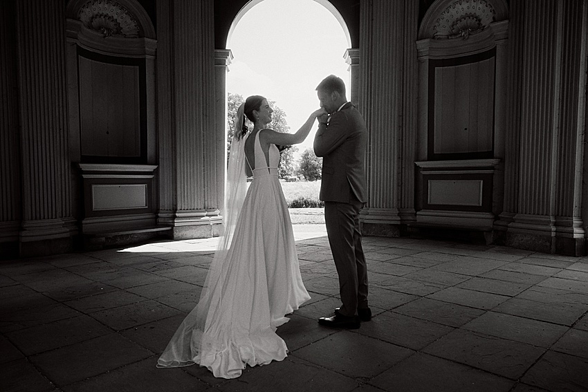 Braut und Bräutigam stehen sich unter einem großen Torbogen in den Herrenhäuser Gärten gegenüber, von hinten vom Sonnenlicht beleuchtet. In einem Moment stiller Interaktion kurz vor ihrer standesamtlichen Hochzeit teilen die Braut in ihrem langen weißen Kleid und der Bräutigam in seinem Anzug eine emotionale Verbindung.