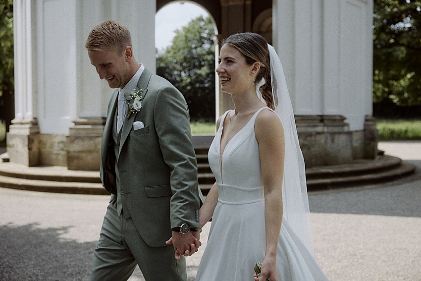 Braut und Bräutigam gehen nach ihrem First Look vor ihrer standesamtlichen Hochzeit Hand in Hand durch das Freie. Der Bräutigam trägt einen hellgrünen Anzug und die Braut ein weißes Kleid mit Schleier. Beide lächeln inmitten der ruhigen Schönheit der Herrenhäuser Gärten in Hannover, eingerahmt von Bäumen und einer Säulenstruktur.