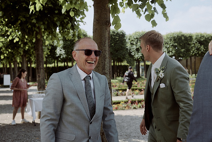 Zwei Männer in Anzügen stehen im Freien und fangen die Freude einer standesamtlichen Hochzeit in der Nähe der Herrenhäuser Gärten Hannover ein. Der Mann links trägt eine Sonnenbrille und einen grauen Anzug und lacht, während der Mann rechts, in einem grünen Anzug mit einer Blumenboutonniere, inmitten von Bäumen und Blumen von der Kamera wegschaut.