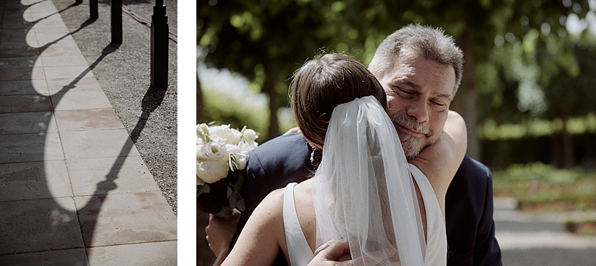 Eine Braut mit Schleier umarmt einen Mann im Anzug im Freien unter Grünpflanzen und feiert ihre standesamtliche Hochzeit. Sie hält einen Strauß weißer Blumen. Links werfen Schatten von Säulen und Lampen komplizierte Muster auf den Steinweg der Herrenhäuser Gärten.