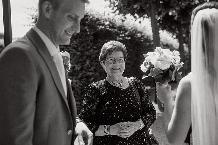 Ein Schwarzweißfoto zeigt eine lächelnde ältere Frau in einem Paillettenkleid mit einer Handtasche bei einer standesamtlichen Hochzeit. Sie steht zwischen einem Mann im Anzug und einer verschleierten Frau mit Blumenstrauß vor der üppigen Kulisse der Herrenhäuser Gärten in Hannover.