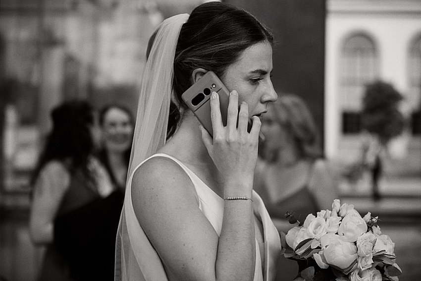 Eine Braut in ärmellosem Kleid und Schleier hält einen Strauß weißer Blumen in einer Hand, während sie bei der Standesamtlichen Hochzeit telefoniert. Sie steht im Freien, umgeben von verschwommenen Gebäuden, und fängt einen zeitlosen Moment in Schwarzweiß ein, der an den klassischen Charme Hannovers erinnert.