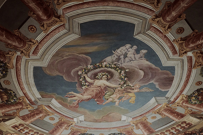 Ein Deckenfresko im Barockstil in den Herrenhäuser Gärten in Hannover zeigt anschaulich eine zentrale Figur inmitten von Wolken, mit einem Engel, der eine Girlande hält. Umgeben von kunstvollen architektonischen Verzierungen und Säulen ist es eine exquisite Wahl für eine elegante Kulisse für eine standesamtliche Hochzeit.