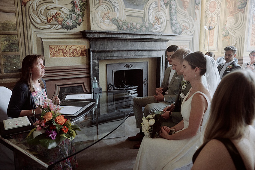 Bei einer standesamtlichen Hochzeit sitzen Braut und Bräutigam Seite an Seite an einem Tisch und sind von der Wärme einer emotionalen Zeremonie umgeben. Im Raum, der mit kunstvollen Wanddekorationen und einem Kamin geschmückt ist, liegt ein Blumenstrauß, während der Geistliche das Paar sanft anspricht.