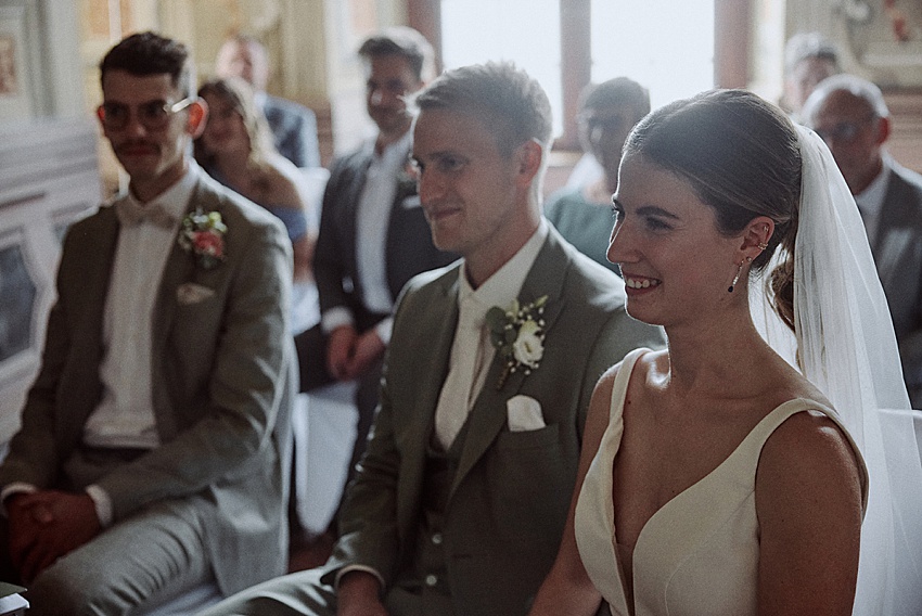 Bei einer standesamtlichen Hochzeit in Hannover sitzen Braut und Bräutigam während der Zeremonie gemeinsam vorne im Raum. Der Bräutigam, in einem hellen Anzug mit Ansteckblume, und die Braut in ihrem weißen Kleid mit Schleier, sind von Gästen umgeben, während sanftes Licht durch die Herrenhäuser Gärten fällt.