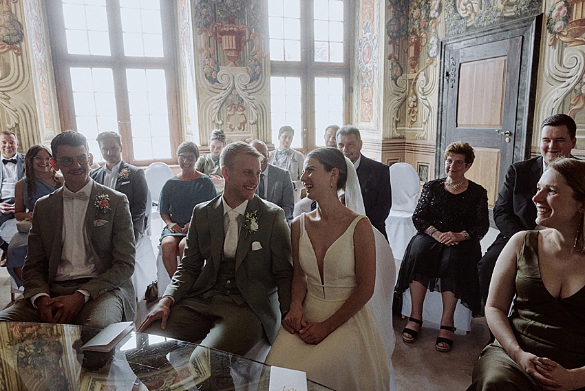 Bei einer standesamtlichen Hochzeit in Hannover sitzt ein Paar in formeller Kleidung lächelnd während der Zeremonie in einem reich verzierten Raum mit großen Fenstern. Um sie herum sitzen die Gäste, ebenfalls formell gekleidet. Der Veranstaltungsort besticht durch detaillierte Wandmalereien und elegante Dekorationselemente, die an die Herrenhäuser Gärten erinnern.