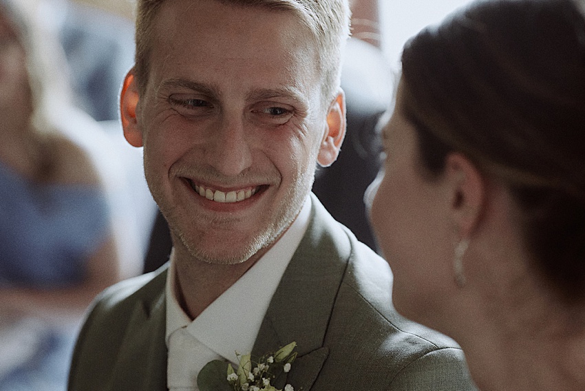 Ein Mann in einem grauen Anzug mit weißer Krawatte und Boutonniere lächelt eine Frau mit hellbraunem Haar während einer scheinbar emotionalen Trauung an. Der leicht unscharfe Hintergrund lässt vermuten, dass die Gäste bei dieser herzlichen standesamtlichen Hochzeit sitzen.