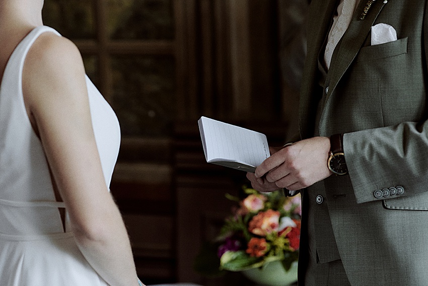 Eine Person im weißen Kleid steht neben einer anderen im grünen Anzug und hält ein Notizbuch in der Hand, möglicherweise bei ihrer standesamtlichen Hochzeit. Sie befinden sich im Innenbereich, mit verschwommenen Blumen im Hintergrund, was an den eleganten Charme der Herrenhäuser Gärten in Hannover erinnert.