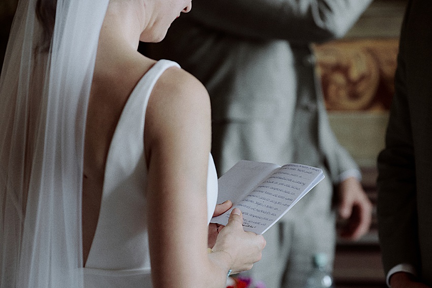 Eine Braut in weißem Kleid und Schleier steht mit dem Rücken zu ihr und hält ein Heft mit gedrucktem Text in der Hand. Im Hintergrund ist teilweise eine Person im Anzug zu sehen. Die Innenkulisse lässt auf einen intimen Moment schließen, möglicherweise während einer standesamtlichen Hochzeit oder einer Probe in den Herrenhäuser Gärten in Hannover.