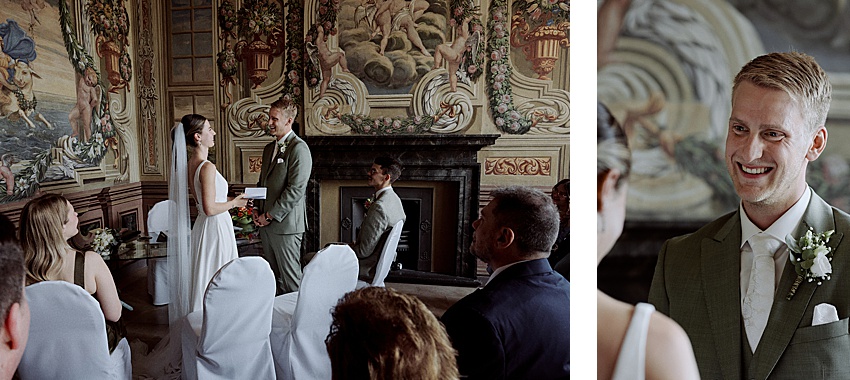 Bei ihrer standesamtlichen Hochzeit stehen sich Braut und Bräutigam in einem kunstvoll verzierten Raum mit detailreichen Wandmalereien gegenüber. Der Bräutigam trägt einen grünen Anzug, während die Braut ein weißes Kleid trägt. Gäste sitzen in der Nähe, einer hält den Moment mit einem Smartphone fest, was an die Eleganz der Herrenhäuser Gärten in Hannover erinnert.