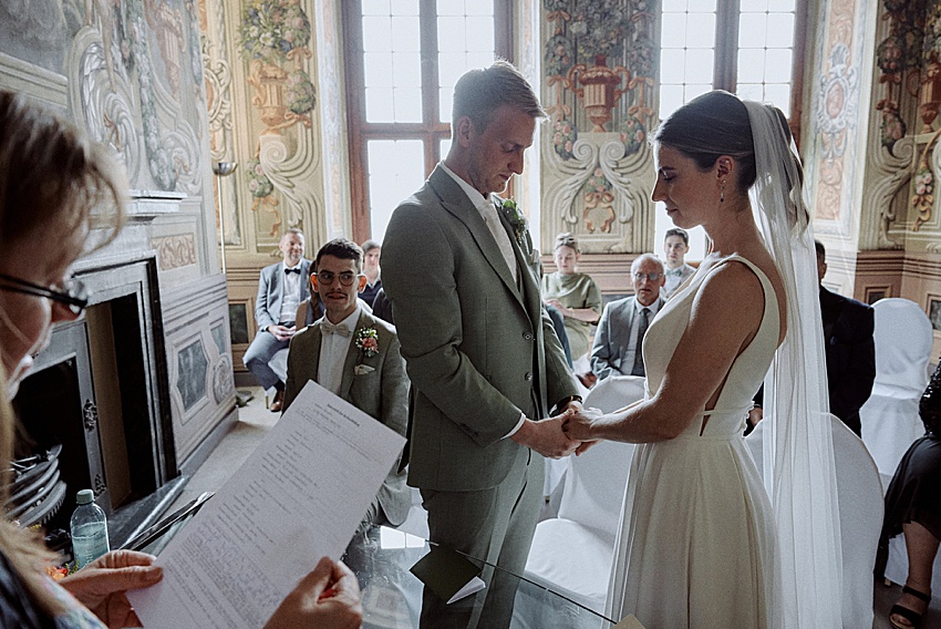 Bei einer standesamtlichen Hochzeit steht ein Paar Händchen haltend in einem reich verzierten Raum mit Blumenmalereien und großen Fenstern, der an die Eleganz der Herrenhäuser Gärten in Hannover erinnert. Der Bräutigam trägt einen hellen Anzug und die Braut ein weißes Kleid mit Schleier. Im Vordergrund hält eine Person ein Blatt Papier, während die Gäste Platz nehmen.