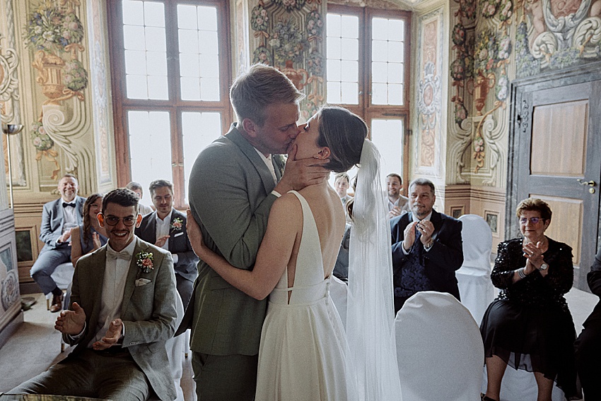 Bei ihrer standesamtlichen Hochzeit in Hannover küssen sich Braut und Bräutigam in einem kunstvoll dekorierten Raum. Die Gäste klatschen und lächeln, während das Sonnenlicht durch große Fenster die Szene erhellt. Das weiße Kleid und der Schleier der Braut ergänzen den hellen Anzug des Bräutigams und erinnern an die Eleganz der Herrenhäuser Gärten.