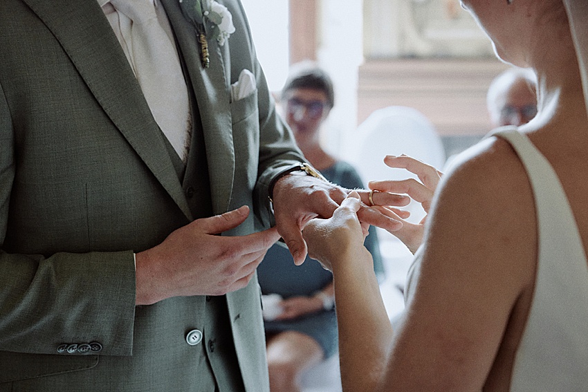 Eine Nahaufnahme einer standesamtlichen Hochzeit zeigt eine Person in einem grauen Anzug und eine Person in einem ärmellosen weißen Kleid, die Ringe austauschen. Im Hintergrund sind zwei sitzende Gäste zu sehen, leicht unscharf, was an eine intime Zeremonie erinnert, die vielleicht in Hannovers romantischen Herrenhäuser Gärten stattfand.