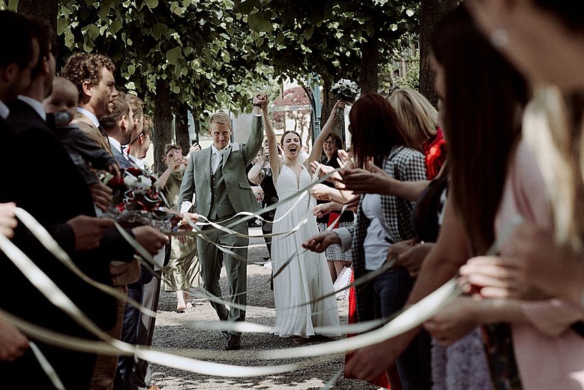 Bei ihrer standesamtlichen Hochzeit in Hannover schlendern Braut und Bräutigam einen von Bäumen gesäumten Weg in den atemberaubenden Herrenhäuser Gärten entlang. Die Braut umklammert ihren Brautstrauß, während die Gäste Bänder hochhalten und einen bezaubernden Torbogen um das frisch vermählte Paar bilden.