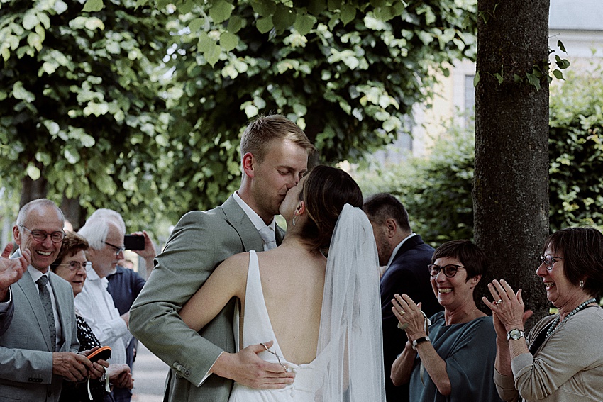 Bei ihrer standesamtlichen Hochzeit in Hannovers malerischen Herrenhäuser Gärten küssen sich Braut und Bräutigam unter den Bäumen. Umgeben von lächelnden und klatschenden Gästen glänzt sie in ihrem weißen Kleid mit Schleier, während er in seinem hellgrauen Anzug schick aussieht und Freude und Feierlichkeit ausstrahlt.