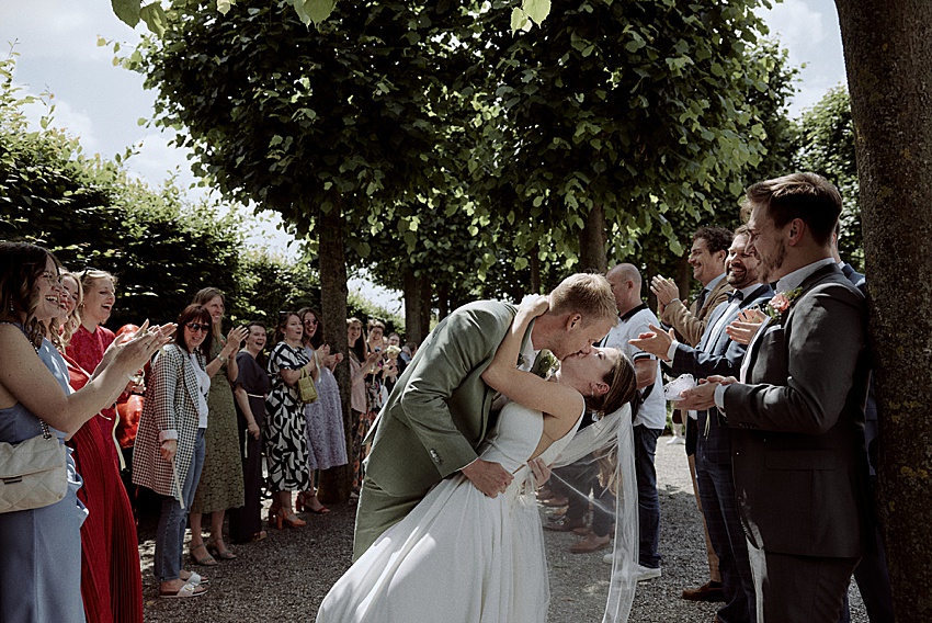 Ein Paar steht bei seiner Standesamtlichen Hochzeit im Freien in leidenschaftlicher Umarmung; der Bräutigam in einem grauen Anzug und die Braut in einem weißen Kleid mit Schleier. Sie sind umgeben von klatschenden Gästen unter dem üppigen grünen Baldachin der Herrenhäuser Gärten in Hannover.