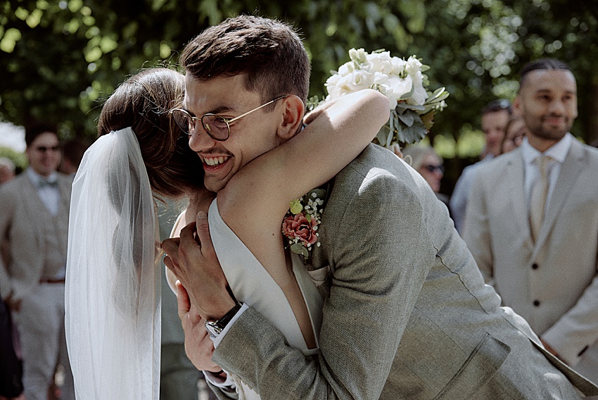 Braut und Bräutigam umarmen sich an ihrem Hochzeitstag im Freien in den malerischen Herrenhäuser Gärten in Hannover. Der Bräutigam, lächelnd in einem hellen Anzug und mit Brille, steht mit seiner Braut zusammen und hält einen Strauß weißer Blumen in der Hand. Gäste in ähnlicher Kleidung sind unter den üppigen grünen Bäumen zu sehen und feiern ihre standesamtliche Hochzeit.