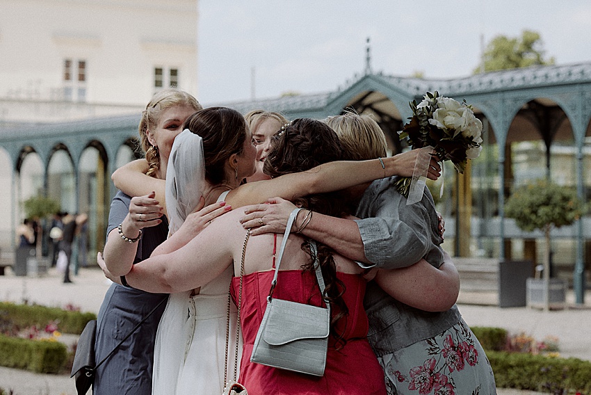 Eine Gruppe von Frauen steht draußen und umarmt sich gegenseitig. Sie strahlen die Freude einer emotionalen Trauung aus. Eine Frau trägt ein weißes Kleid und einen Schleier, was auf eine standesamtliche Hochzeit hindeutet. Ein Blumenstrauß wird gehalten, während die atemberaubende Kulisse der Herrenhäuser Gärten Hannover die wunderschöne Szenerie ergänzt.