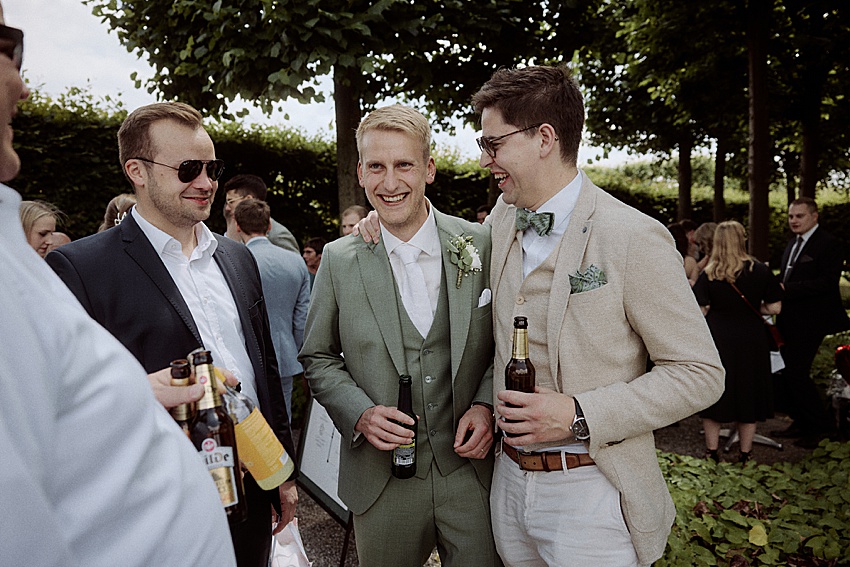 Drei Männer in Anzügen stehen zusammen im Freien und lächeln während einer standesamtlichen Hochzeit in den Herrenhäuser Gärten in Hannover. Der Mann in der Mitte trägt einen grünen Anzug mit einer Ansteckblume, während die anderen beiden beige und dunkle Anzüge tragen. Sie halten Bierflaschen, umgeben von Bäumen und anderen Gästen.