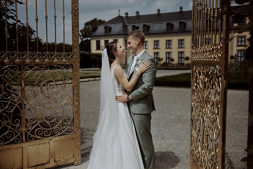 Eine Braut in weißem Kleid und Schleier und ein Bräutigam in hellem Anzug stehen vor einem verzierten goldenen Tor und lächeln einander an. Ihre standesamtliche Hochzeit findet vor der Kulisse des großen Gebäudes und der gepflegten Gärten der Herrenhäuser Gärten unter einem bewölkten Himmel statt.