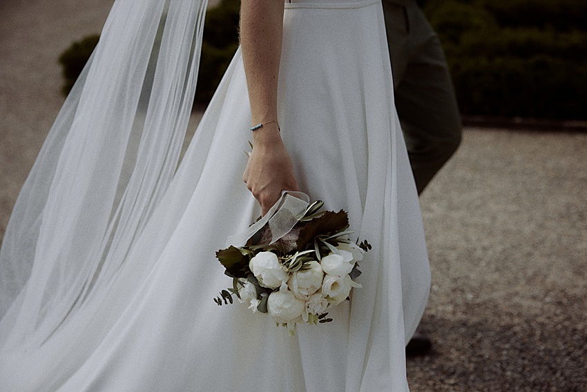 Eine Person in einem weißen Brautkleid mit langem Schleier hält einen Strauß weißer Blumen und Grünpflanzen in den Händen. Die Szene spielt sich draußen auf einem Kiesweg in den Herrenhäuser Gärten Hannover ab, perfekt für eine emotionale Trauung, wobei verschwommenes Grün die ideale Kulisse bietet.