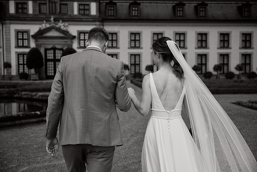 Braut und Bräutigam gehen Hand in Hand vor einem eleganten Gebäude mit großen Fenstern und einem Innenhof, der vielleicht an die majestätischen Herrenhäuser Gärten erinnert. Der Bräutigam trägt einen Anzug und die Braut ein langes Kleid mit Schleier. Das Bild ist wunderschön in Schwarzweiß aufgenommen.