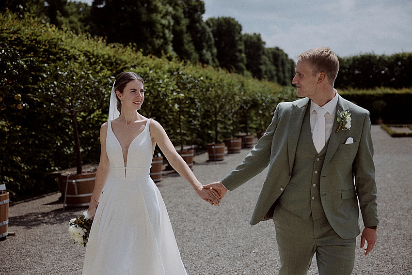 Braut und Bräutigam halten sich auf einem Kiesweg in den malerischen Herrenhäuser Gärten Hannover an den Händen. Die Braut trägt ein weißes Kleid und einen Schleier und hält einen Blumenstrauß in der Hand, während der Bräutigam einen grünen Anzug trägt. Grüne Büsche und Bäume bilden unter einem leicht bewölkten Himmel die Kulisse für diese emotionale Trauung.