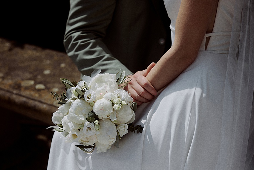 Eine Braut in einem weißen Kleid sitzt neben einer Person in einem dunklen Outfit und hält einen Strauß weißer Blumen in der Hand. Während sie sich an den Händen halten, lässt der intime Moment auf eine emotionale Trauung schließen, die vielleicht in den malerischen Herrenhäuser Gärten Hannover eingefangen wurde und auf eine romantische oder standesamtliche Hochzeit schließen lässt.