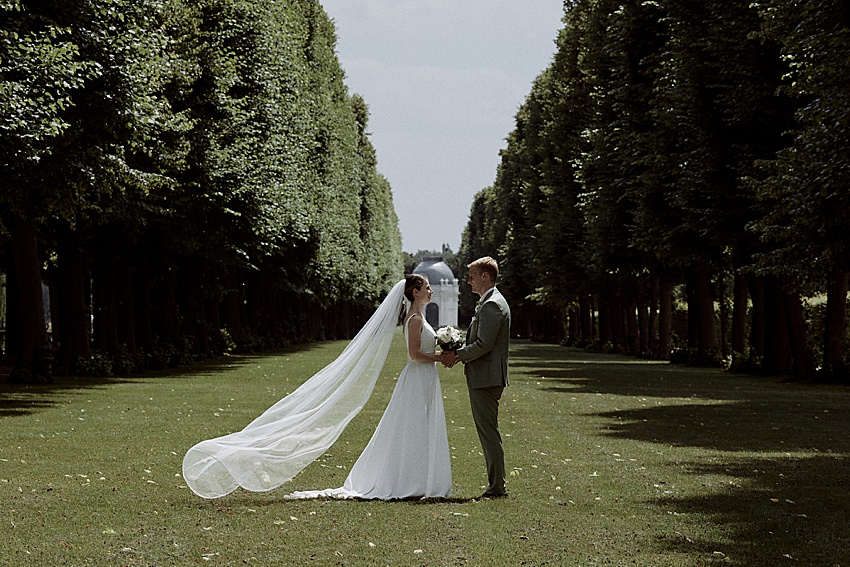 Braut und Bräutigam stehen sich auf einem von Bäumen gesäumten Weg in den Herrenhäuser Gärten in Hannover gegenüber. Die Braut in weißem Kleid und Schleier hält einen Blumenstrauß, während der Bräutigam einen hellen Anzug trägt. Der lange Schleier fließt zur Seite, eingerahmt von hohen Bäumen, die eine symmetrische Kulisse für ihre Standesamtliche Hochzeit bilden.