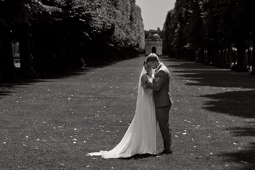 Braut und Bräutigam umarmen sich in einem von Bäumen gesäumten Park in den Herrenhäuser Gärten und feiern ihre standesamtliche Hochzeit. Die Braut trägt ein langes weißes Kleid und der Bräutigam einen Anzug. Verstreute Blütenblätter bedecken den Weg, während ein entferntes Gebäude den Hintergrund der schwarz-weißen Szene in Hannover ziert.