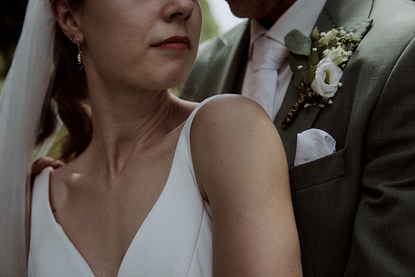 Nahaufnahme einer Braut in einem weißen Kleid und Schleier, die ihre Schulter und einen Teil ihres Gesichts zeigt, neben einem Bräutigam in einem grünen Anzug mit einer Blumenboutonniere. Das Bild fängt einen intimen Moment mit natürlichem Licht während ihrer standesamtlichen Hochzeit in den eleganten Herrenhäuser Gärten Hannover ein.