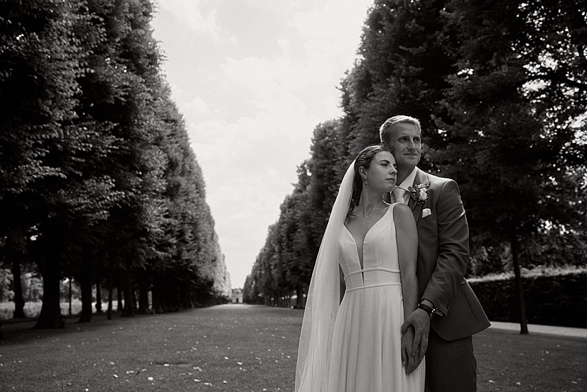 Ein Schwarzweißfoto zeigt ein Paar bei seiner Standesamtlichen Hochzeit in Hannover. Sie umarmen sich eng, eingerahmt von dem von Bäumen gesäumten Weg der Herrenhäuser Gärten, der sich in die Ferne erstreckt. Die Braut trägt Schleier und Kleid, während der Bräutigam einen Anzug mit Ansteckblume trägt.