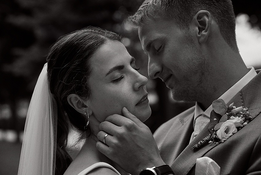 Ein Schwarzweißbild zeigt ein Paar, das sich während seiner Standesamtlichen Hochzeit umarmt. Die Frau trägt einen Schleier und hat in aller Gelassenheit die Augen geschlossen. Der Mann, der an seinem Anzug eine Ansteckblume trägt, berührt sanft ihr Gesicht. Ihr liebevoller Moment spiegelt die Eleganz der Herrenhäuser Gärten in Hannover wider.