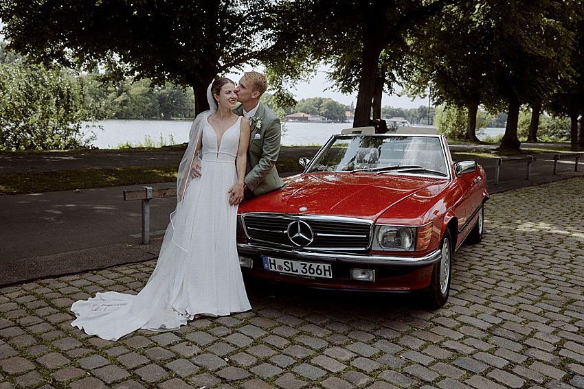 Ein frisch vermähltes Paar steht neben einem roten Mercedes-Oldtimer-Cabrio auf einem Kopfsteinpflasterweg in den Herrenhäuser Gärten. Die Braut trägt ein weißes Kleid und einen Schleier, während der Bräutigam einen hellgrünen Anzug trägt. Im Hintergrund sind Bäume und ein See zu sehen.