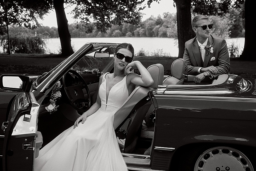 Eine Braut im weißen Kleid sitzt in einem klassischen Cabrio mit offener Tür und trägt eine Sonnenbrille. Ihr Bräutigam, ebenfalls mit Sonnenbrille, lehnt an der Seite des Autos. Dieser elegante Moment in den Herrenhäuser Gärten, in Schwarzweiß festgehalten, fängt ihre standesamtliche Hochzeit am ruhigen Wasser von Hannover perfekt ein.