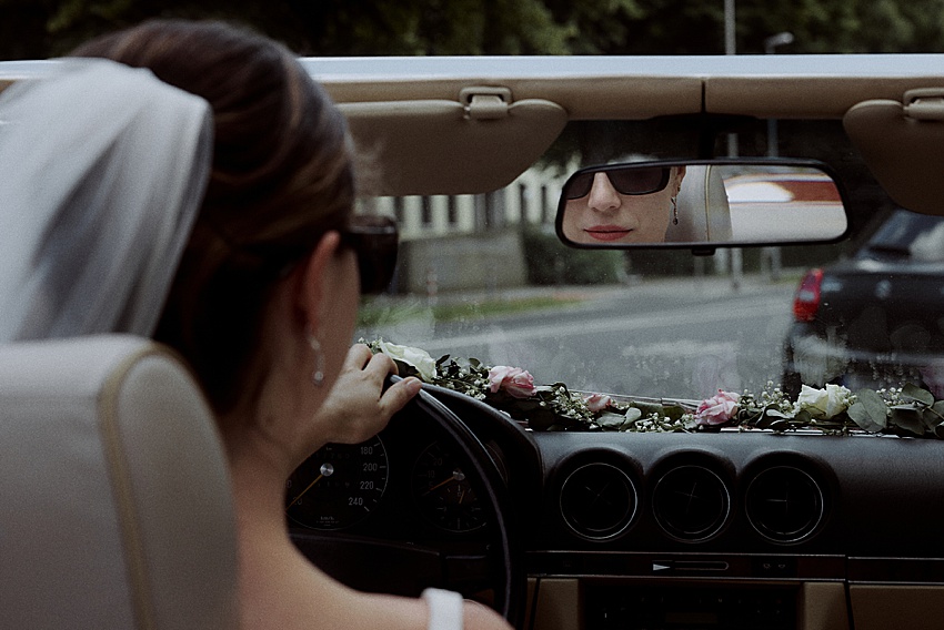 Eine Braut mit Schleier fährt mit einem Cabrio zu ihrer Standesamtlichen Hochzeit in Hannover. Rosa und weiße Blumen schmücken das Armaturenbrett und ihr Spiegelbild schimmert im Rückspiegel, während sie an anderen Fahrzeugen auf der Straße vorbeifährt und von Feierlichkeiten in den Herrenhäuser Gärten träumt.